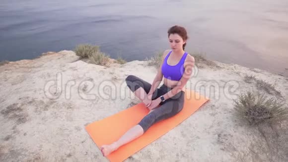可爱的运动白种人女孩坐在海洋上方的悬崖边做伸展运动日落时的户外运动视频的预览图