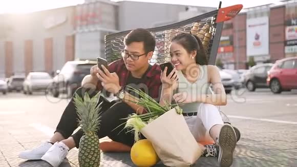 可爱的亚洲青年男女穿着休闲服装坐在超市前的地上看着他们的表演视频的预览图