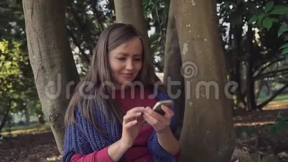 可爱的女孩坐在公园的树旁她用智能手机微笑着视频的预览图