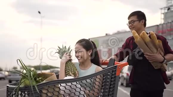可爱的年轻亚洲女人坐在购物车里手里拿着纸袋把她英俊的男朋友推着吃东西视频的预览图