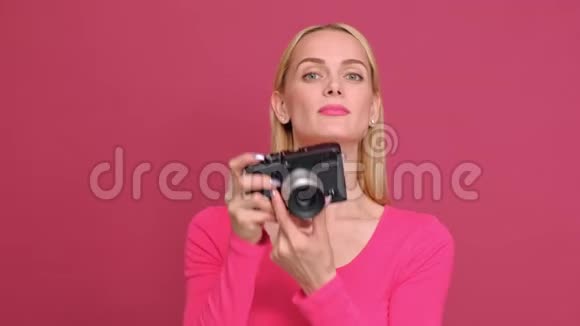 漂亮的年轻女子金发穿着红色T恤和牛仔裤摆在粉红色的背景上无镜相机上的照片视频的预览图