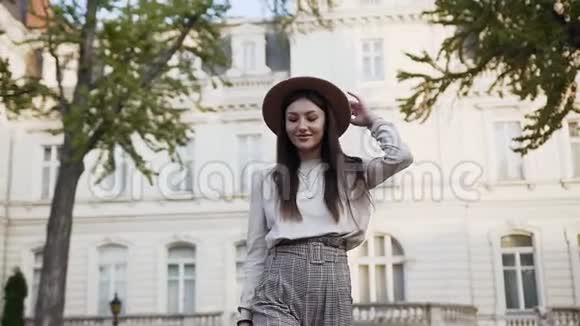 阳光明媚的一天漂亮的年轻女子戴着时髦的帽子走在豪华的豪宅附近视频的预览图