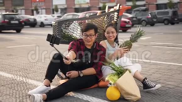 满意的年轻亚洲夫妇坐在市场手推车附近的地上用苹果自拍视频的预览图