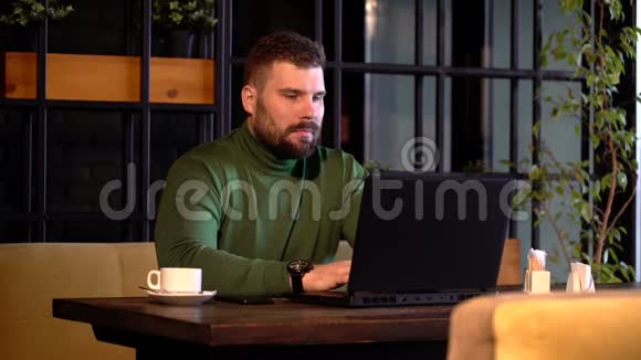 年轻有魅力的胡子男在咖啡厅休息时自信地工作在笔记本电脑上视频的预览图