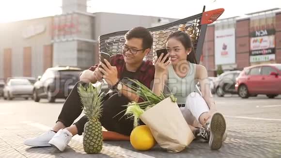 快乐漂亮的亚洲男人和女人坐在市场手推车附近的沥青上用他们的手机看有趣的节目视频的预览图