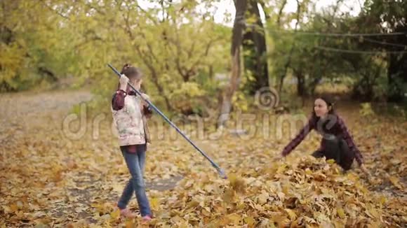 美丽的小女孩和她的母亲用耙子清理院子里的黄叶视频的预览图