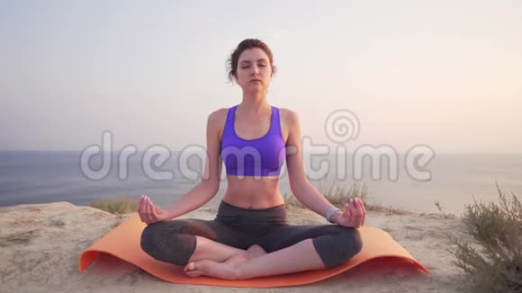 一个年轻而有魅力的女人坐在莲花的位置上专注于呼吸黎明时在山上的海边视频的预览图
