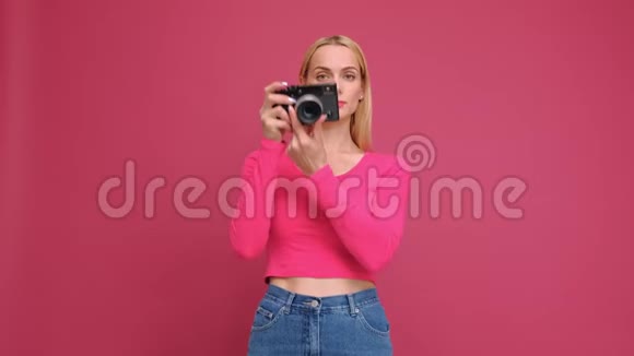 漂亮的年轻女子金发穿着红色T恤和牛仔裤摆在粉红色的背景上无镜相机上的照片视频的预览图