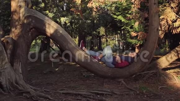 一个可爱的女孩躺在公园的一棵大树的半圆形树干上半圆形树干上休息视频的预览图