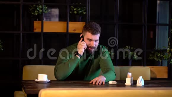 年轻的大胡子开朗的男人坐在桌旁的咖啡馆里在电脑上工作在手机上聊天视频的预览图