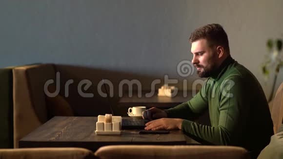 大胡子的时髦商人坐在桌子旁的咖啡馆里用笔记本电脑工作用手机说话视频的预览图