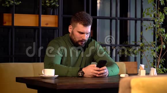 年轻的留胡子的时髦男人坐在咖啡馆的桌子旁用智能手机喝咖啡视频的预览图