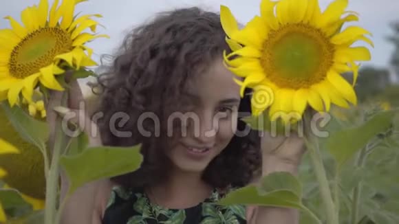 一幅可爱的卷发女孩看着镜头微笑着用向日葵田野上的两朵向日葵遮住她的脸的肖像视频的预览图
