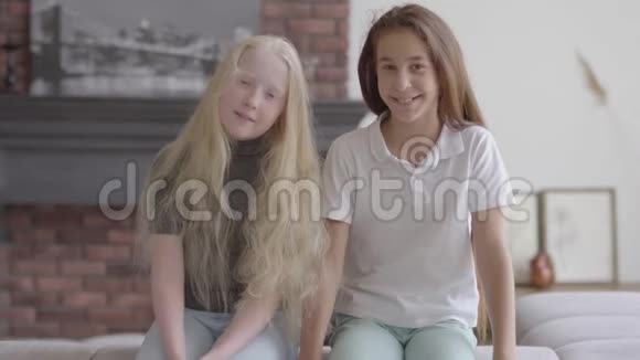 两个可爱的微笑女孩坐在沙发上看着客厅里的摄像机友谊的概念免费视频的预览图