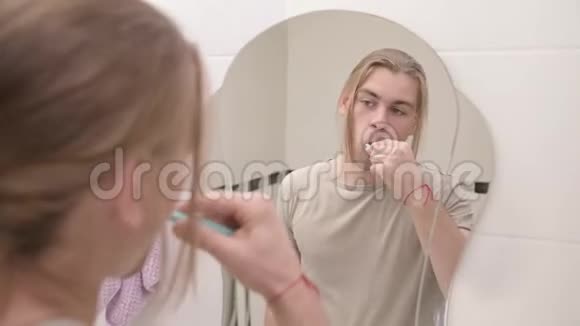 年轻迷人的千禧一代长发男子保持早晨的口腔卫生在浴室里用镜子刷牙视频的预览图