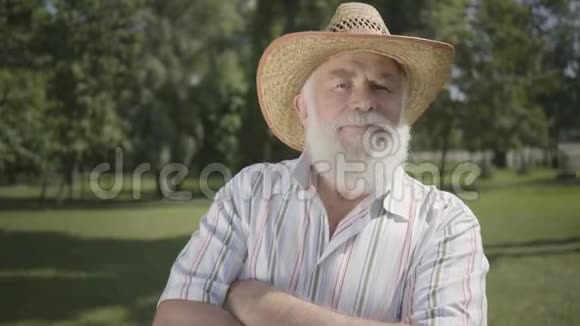 自信的灰色胡子老人戴着草帽看着摄像机微笑地站在公园里户外休闲成熟的男人视频的预览图