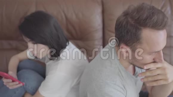 特写一幅悲伤的男人和女人背靠背坐在家里脸上带着不快乐的表情那个用手机的女人视频的预览图