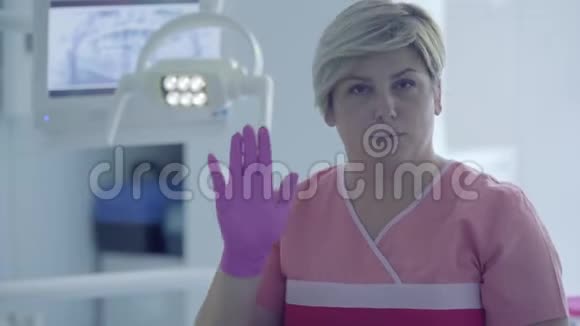 在牙科办公室的工作场所戴着医用手套的可爱的女牙医在看摄像机时显示出手势女性视频的预览图