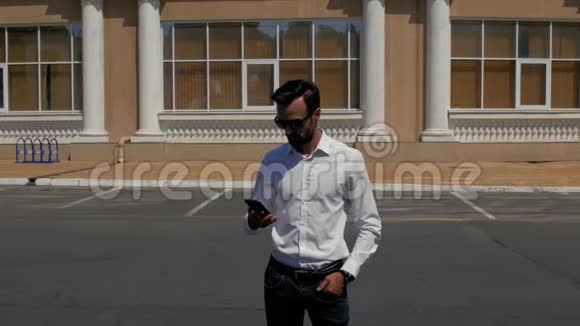 时尚的年轻人留着胡子穿着白色衬衫戴着太阳镜站在一座现代化的建筑前打电话4k视频的预览图