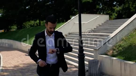 一个穿着夹克和白衬衫留着胡子的商人走在路上的公园里看着他手上的手表走了视频的预览图