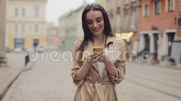美丽的少女戴着耳机在她的智能手机发短信笑站在城市背景视频的预览图