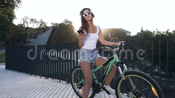 美丽的热辣的微笑的年轻女子戴着时髦的眼镜坐在自行车上在城市里听着耳机里的音乐视频的预览图