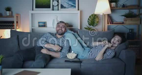 欢乐的男人和女人看电视晚上在家里笑着扔爆米花视频的预览图