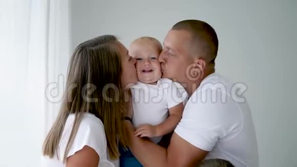 爸爸和妈妈穿着白色t恤亲吻一个一岁的孩子的脸颊视频的预览图
