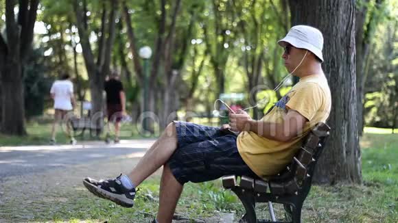 戴帽子戴太阳镜的人坐在公园的长凳上听音乐男人一边听音乐一边在爸爸家放松视频的预览图