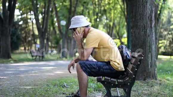 戴着帽子和太阳镜的人坐在公园长凳上打电话在树下的公园里放松时男人用电话交谈视频的预览图