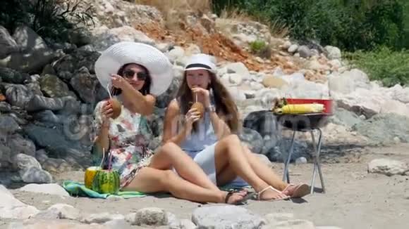 年轻漂亮的女孩戴着帽子戴着墨镜一边喝着新鲜的椰子鸡尾酒一边在沙滩上谈笑风生视频的预览图
