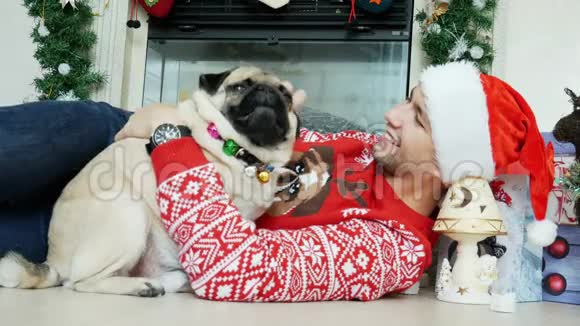 穿圣诞服装的男人拥抱和抚摸滑稽的小狗戴着圣诞帽背景中有礼物视频的预览图