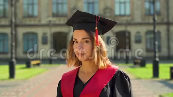 戴着黑色斗篷和帽子的漂亮女孩站在大学大楼前看镜头的肖像青年视频的预览图