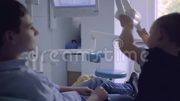 一个十几岁的男孩坐在牙医办公室的椅子上他的弟弟在模仿医生玩台灯孩子们视频的预览图