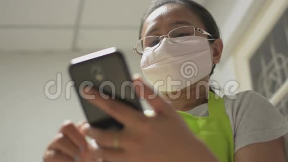 家庭主妇戴着防护面罩使用手机智能手机进行交流并在家里在线观看社交视频的预览图