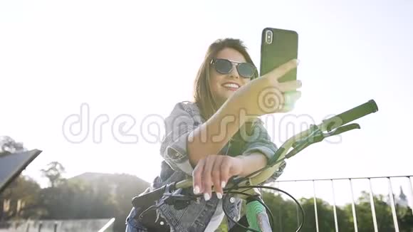 漂亮快乐的女孩戴着时髦的太阳镜和时髦的衣服坐在自行车上在阳光下做有趣的自拍视频的预览图