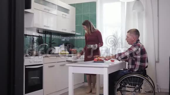 快乐的一对夫妻坐在轮椅上的快乐男人在厨房做饭的时候和妻子玩得很开心视频的预览图