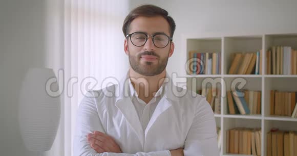 穿着白大褂戴着眼镜看着图书馆里的摄像机的成年白种人英俊男医生的特写照片视频的预览图