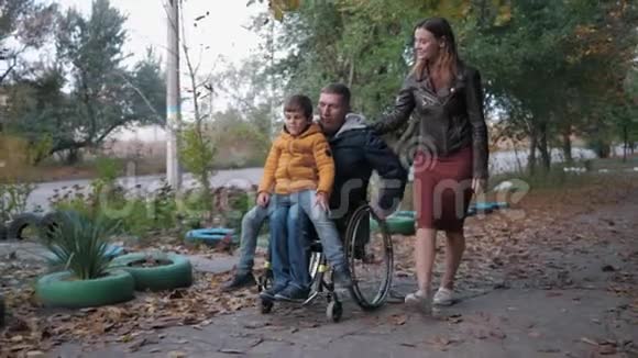 家庭散步残疾的父亲坐在轮椅上抱着儿子跪在秋日街上和他微笑的妻子一起散步视频的预览图