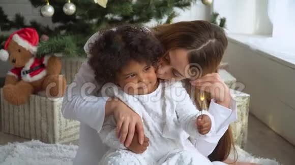 快乐的母亲和穿着白色衣服的小女儿坐在圣诞树旁手里拿着火花视频的预览图