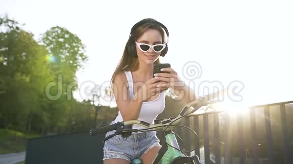 靠近微笑的年轻可爱的女士戴着耳机坐在自行车上听音乐视频的预览图