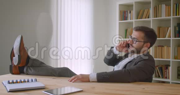 一位英俊的白种人成年商人的特写照片他的脚踩在桌子上打电话视频的预览图