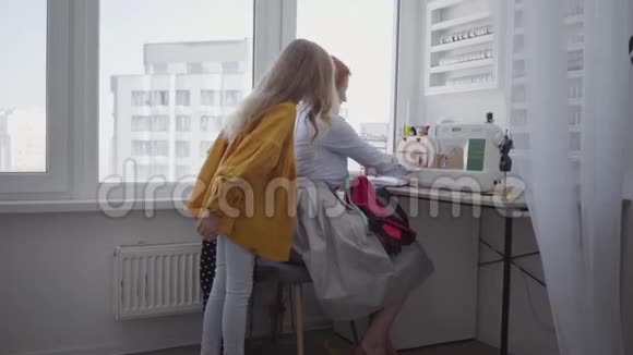 美丽的红发女人缝衣服坐在桌子旁边的窗户在灯光室小女儿看上去视频的预览图