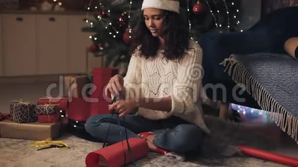 戴着圣诞帽准备圣诞礼物缠着绷带系着蝴蝶结坐在圣诞树下的漂亮女孩视频的预览图