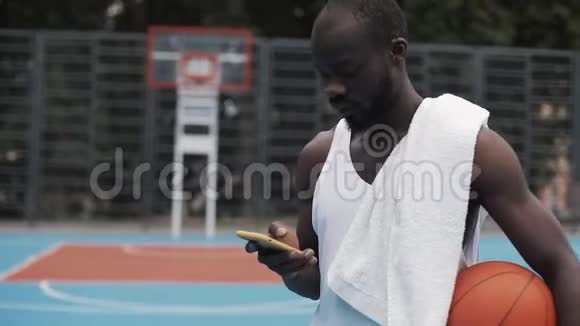 一个年轻的肌肉发达的非裔美国人穿着白色单打拿着球用他的智能手机打信息视频的预览图