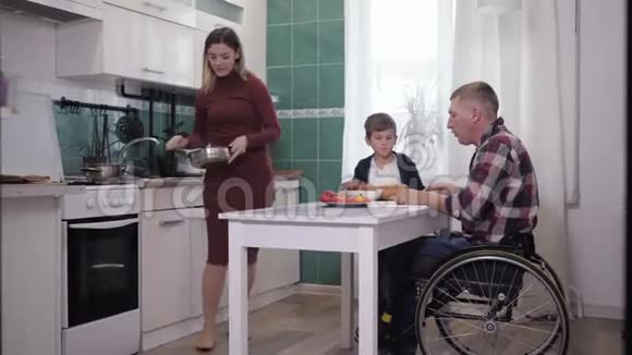 家庭互动一个坐在轮椅上的男人和他的妻子和儿子一起在厨房准备食物好好说话好好说话视频的预览图