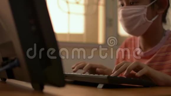 可爱的女孩戴着防护面罩在COVID19疫情爆发期间在家里的桌子上使用电脑视频的预览图