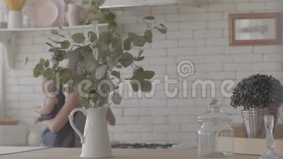 年轻的母亲在厨房里准备食物把婴儿抱在怀里有绿叶的花瓶视频的预览图