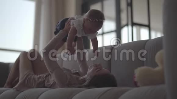 美丽苗条的年轻女子躺在沙发上把她的女婴抬起来母亲在家里和孩子玩耍视频的预览图