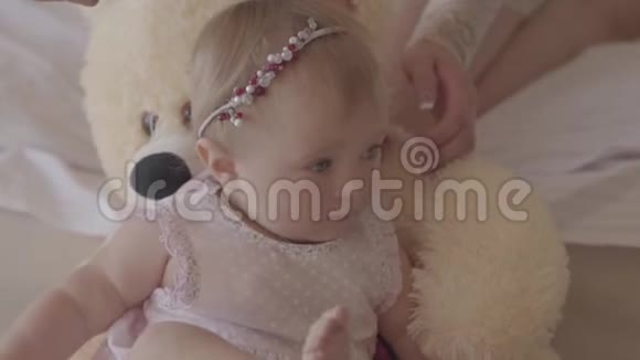一个穿着粉红色连衣裙的小女孩躺在家里的熊玩具上而她母亲的手在她的头发上刷头发视频的预览图